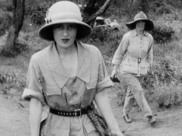 Two women dressed in safari-style outfits and wide-brimmed hats walk outdoors on a dirt path surrounded by vegetation in a black-and-white, vintage photograph.