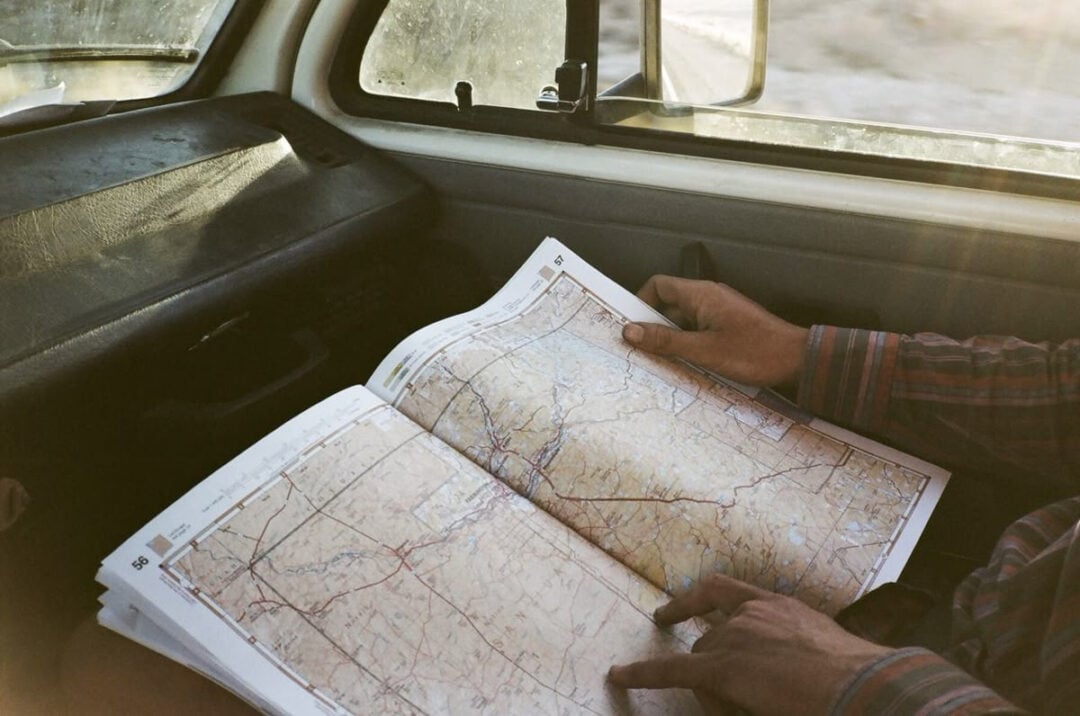 A person sitting in a car holds an open road map, studying it as sunlight streams through the window onto the dashboard and their hands.