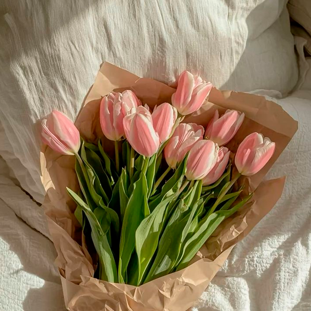 A bouquet of pink tulips with green leaves is wrapped in brown paper and placed on a soft, white, ruffled bedspread in natural sunlight.