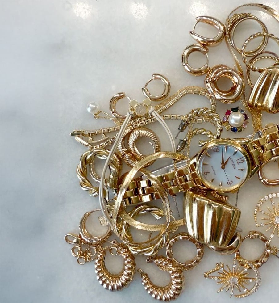 A pile of gold jewelry, including bracelets, rings, chains, and a wristwatch, is arranged on a white marble surface. The items are shiny and intricately designed, with some featuring gemstones and decorative patterns.