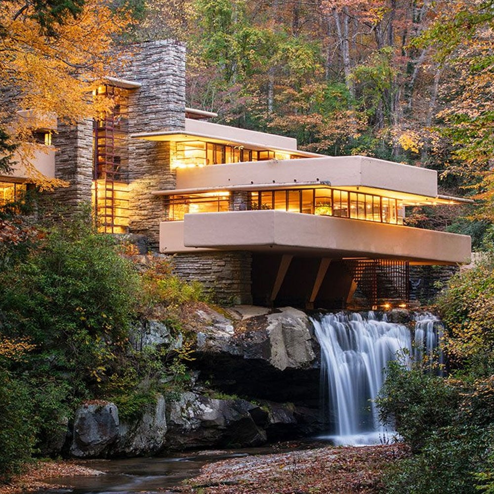 A modern house with stone and concrete features is built over a waterfall, surrounded by autumn trees with orange and yellow leaves, blending into the forest landscape.