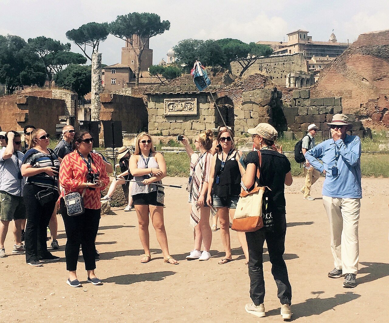A tour guide speaks to a group of tourists at an ancient historic site with stone ruins, trees, and old buildings in the background. The group listens attentively, some wearing sunglasses and holding cameras.