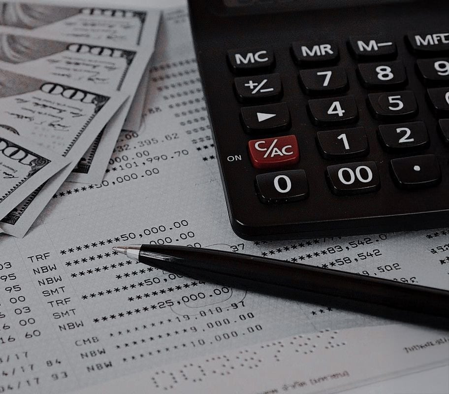 A close-up of a calculator, a pen, several $100 bills, and a bank statement with transactions and account numbers visible, suggesting financial calculations or budgeting.