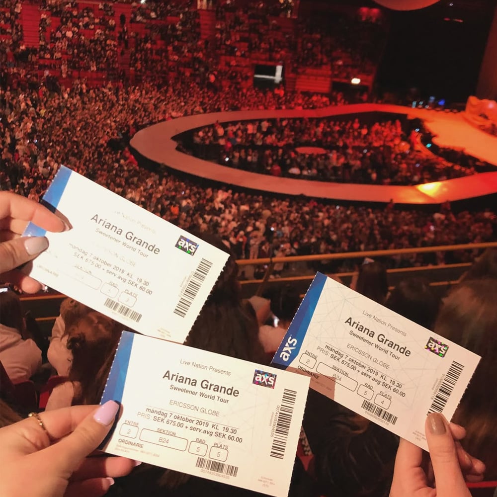 Three people hold up Ariana Grande concert tickets inside a large, crowded arena. The stage is visible in the background, with fans filling the seats and anticipation in the air before the show begins.