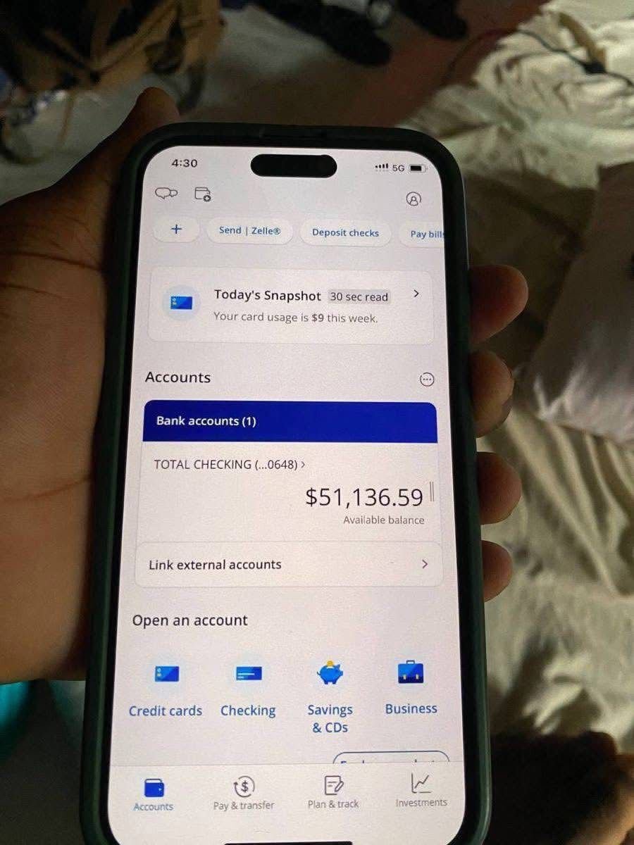 A hand holding a smartphone displaying a banking app screen, showing a checking account with a balance of $51,136.59. In the background, a messy bed and floor are partially visible.