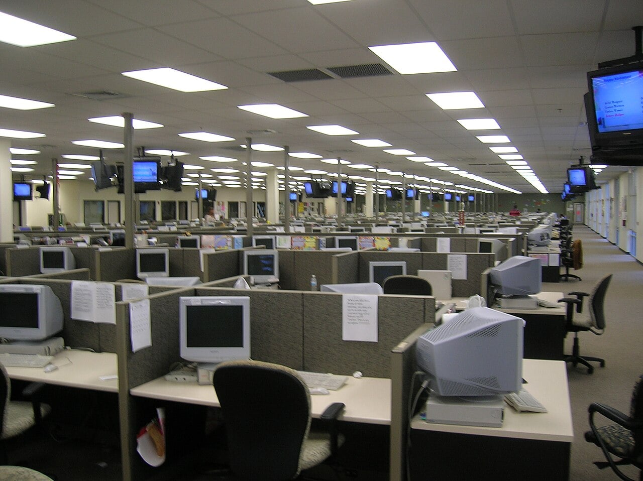 A large, empty office space with many cubicles, each containing a computer, chair, and paperwork. Several monitors hang from the ceiling throughout the room. The area is brightly lit with fluorescent lights.