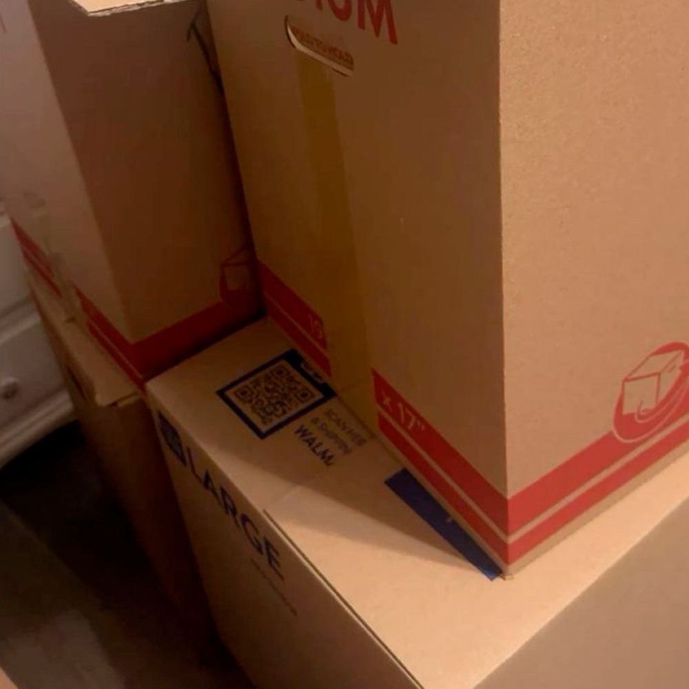 Several packed cardboard moving boxes are stacked on top of each other in a dimly lit room. Some boxes have red stripes and labels, and one box is marked "LARGE" with a visible QR code.