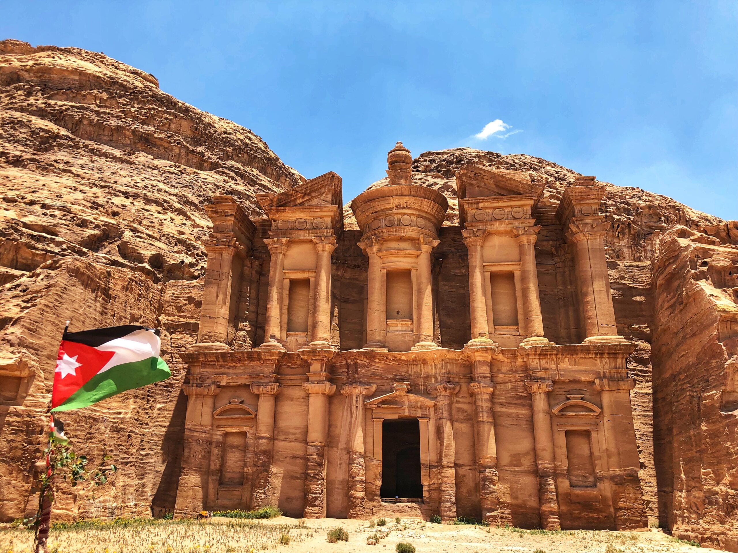 Ancient stone facade of the Monastery (Ad Deir) in Petra, Jordan, stands against rocky hills under a blue sky, with the Jordanian flag visible in the foreground.