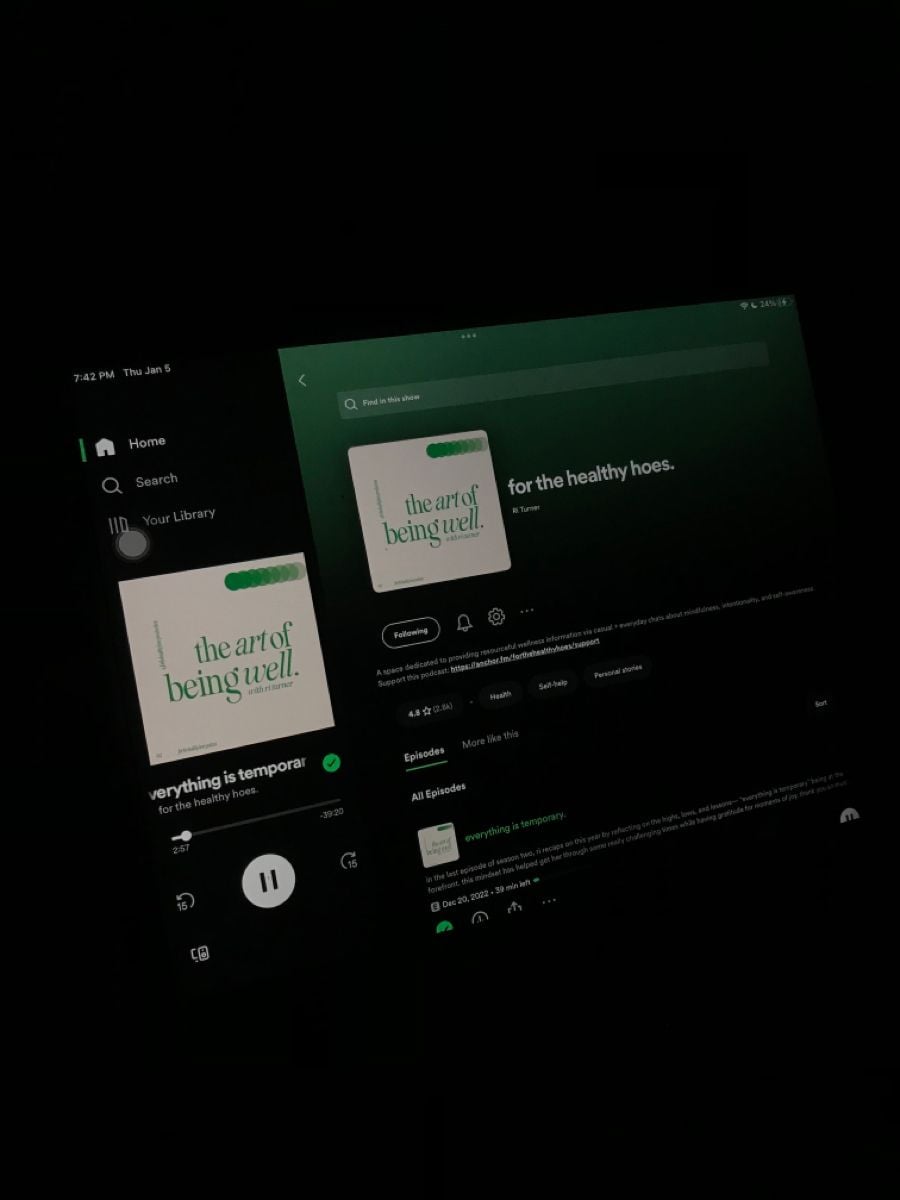 A dark room with a tablet and a phone screen displaying the podcast "the art of being well" on Spotify. Both screens show the podcast’s cover and episode details with dark backgrounds.