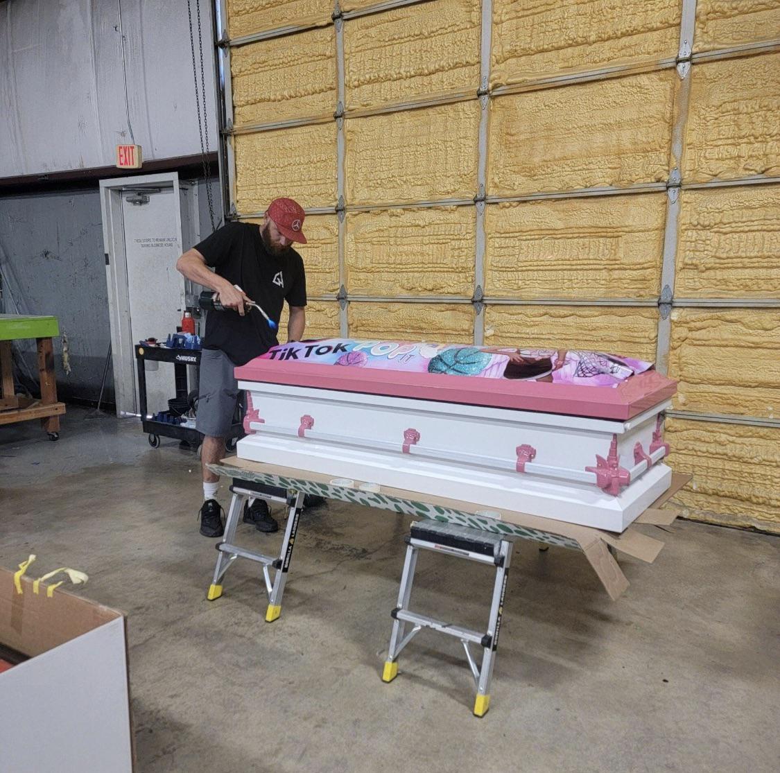 A man wearing a red cap and black shirt works on painting a pink and white coffin decorated with "TikTok" text and colorful designs in a large workshop.