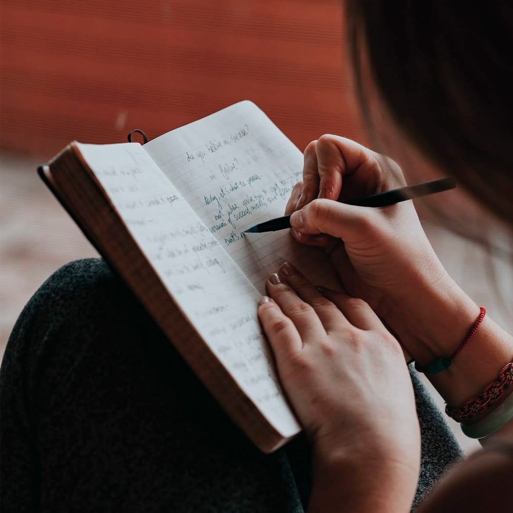 A person sitting with crossed legs writes in a notebook using a black pen. The notebook contains handwritten notes, and the person wears a thin red bracelet on their wrist.