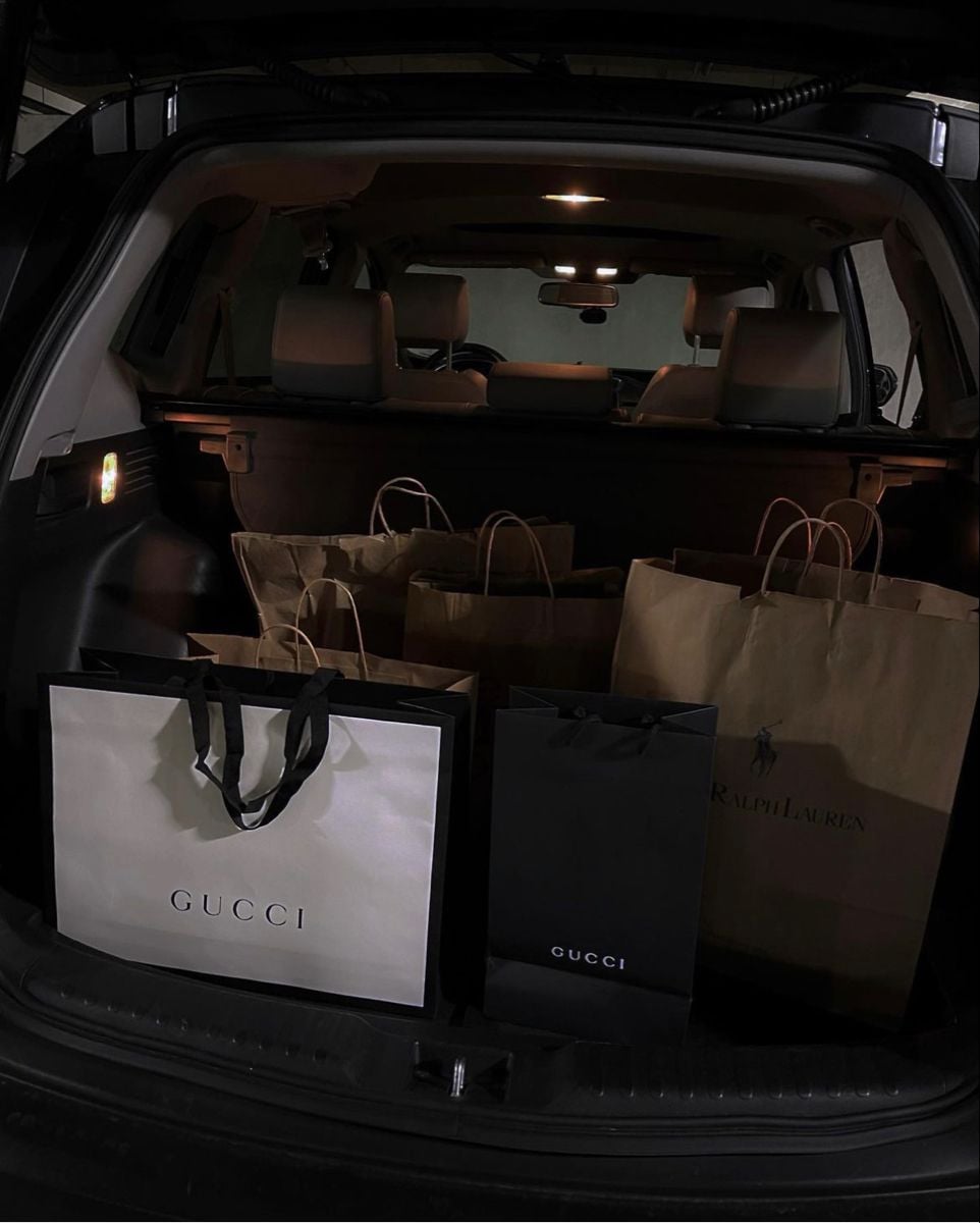 Several Gucci and Ralph Lauren shopping bags are placed in the trunk of a car, with the back seats and interior dimly lit.