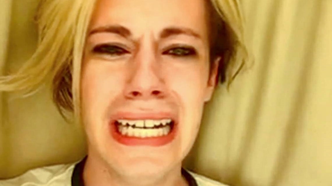 A person with blonde hair and heavy eye makeup is crying intensely, mouth open and eyes red with tears, against a beige background.