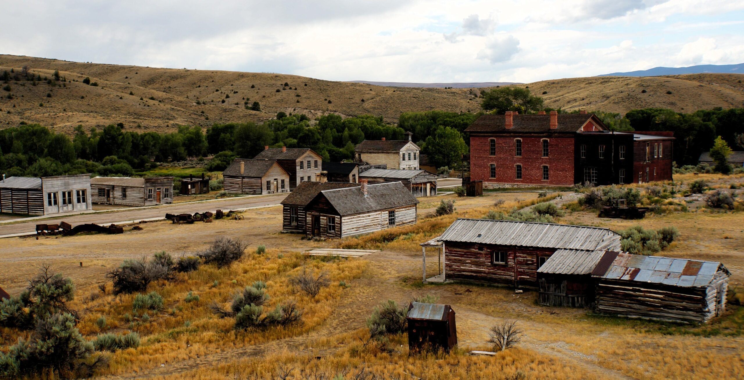 An old western ghost town with wooden and brick buildings scattered across dry, grassy land, surrounded by hills and sparse vegetation under a cloudy sky.
