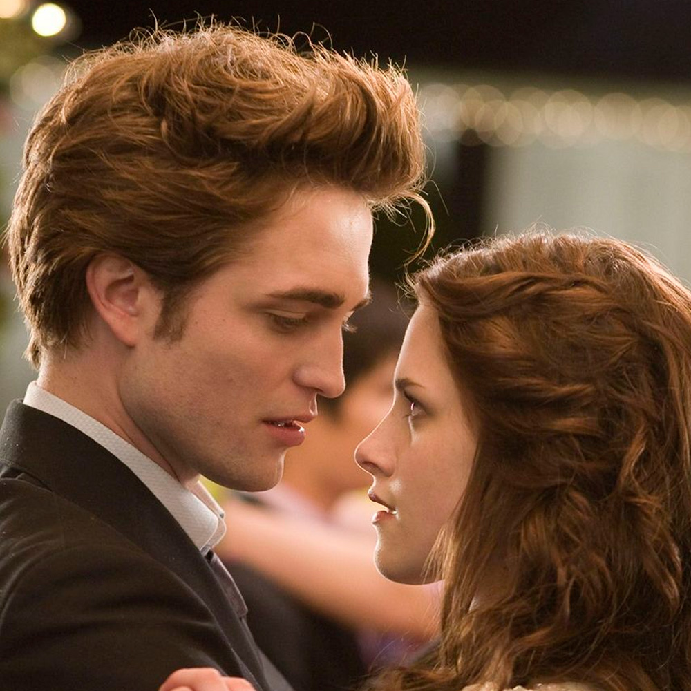 A young man and woman stand closely, gazing into each other's eyes while dancing. The background is softly blurred with lights, giving a romantic atmosphere. Both have serious, intense expressions.