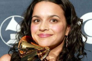 A woman with long dark hair smiles while holding a gold Grammy award trophy. A backdrop with the Grammy logo and CBS eye logo is visible behind her.