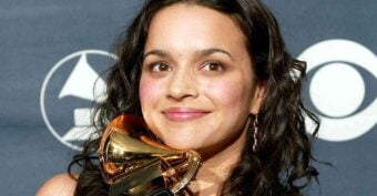 A woman with long dark hair smiles while holding a gold Grammy award trophy. A backdrop with the Grammy logo and CBS eye logo is visible behind her.