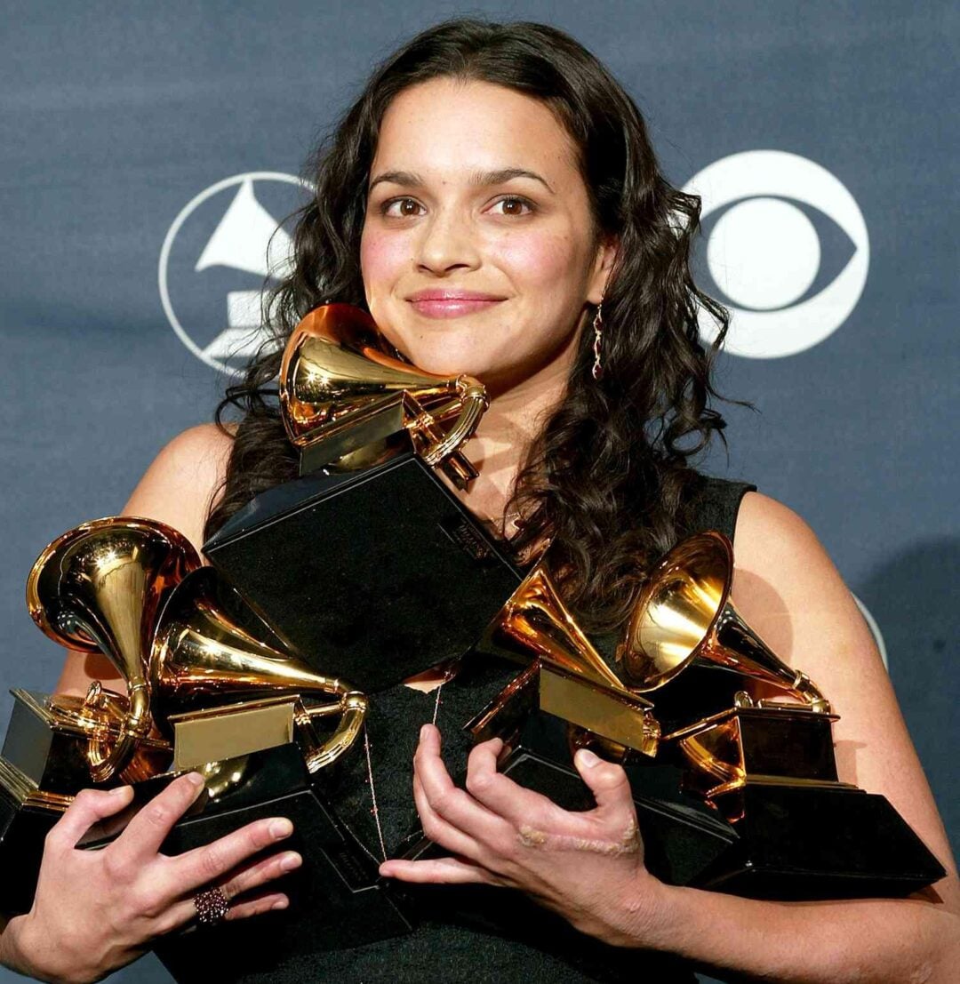 A woman with long, dark hair smiles while holding multiple Grammy Awards in her arms, standing in front of a backdrop with CBS and Grammy logos.