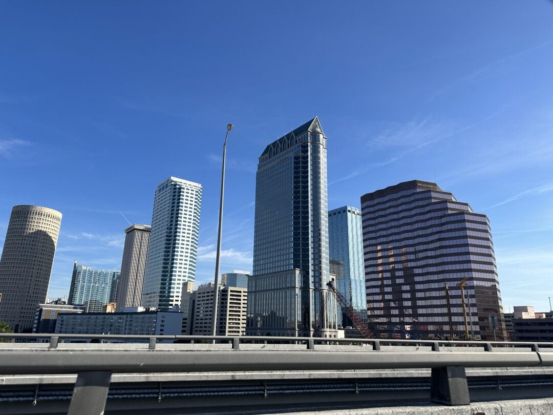 View of several modern skyscrapers and office buildings under a clear blue sky, seen from behind a highway guardrail in an urban cityscape.