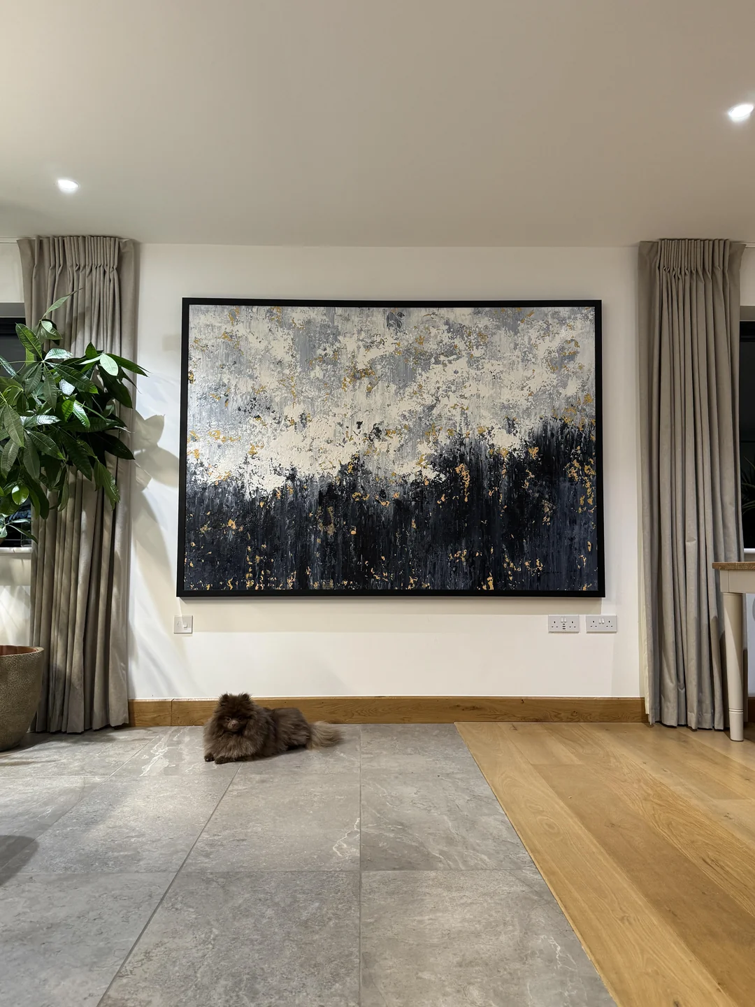 A fluffy dark cat lies on a gray tile floor in front of a large, abstract painting with black, white, and gold colors. The room has light walls, long gray curtains, and a potted plant to the left.