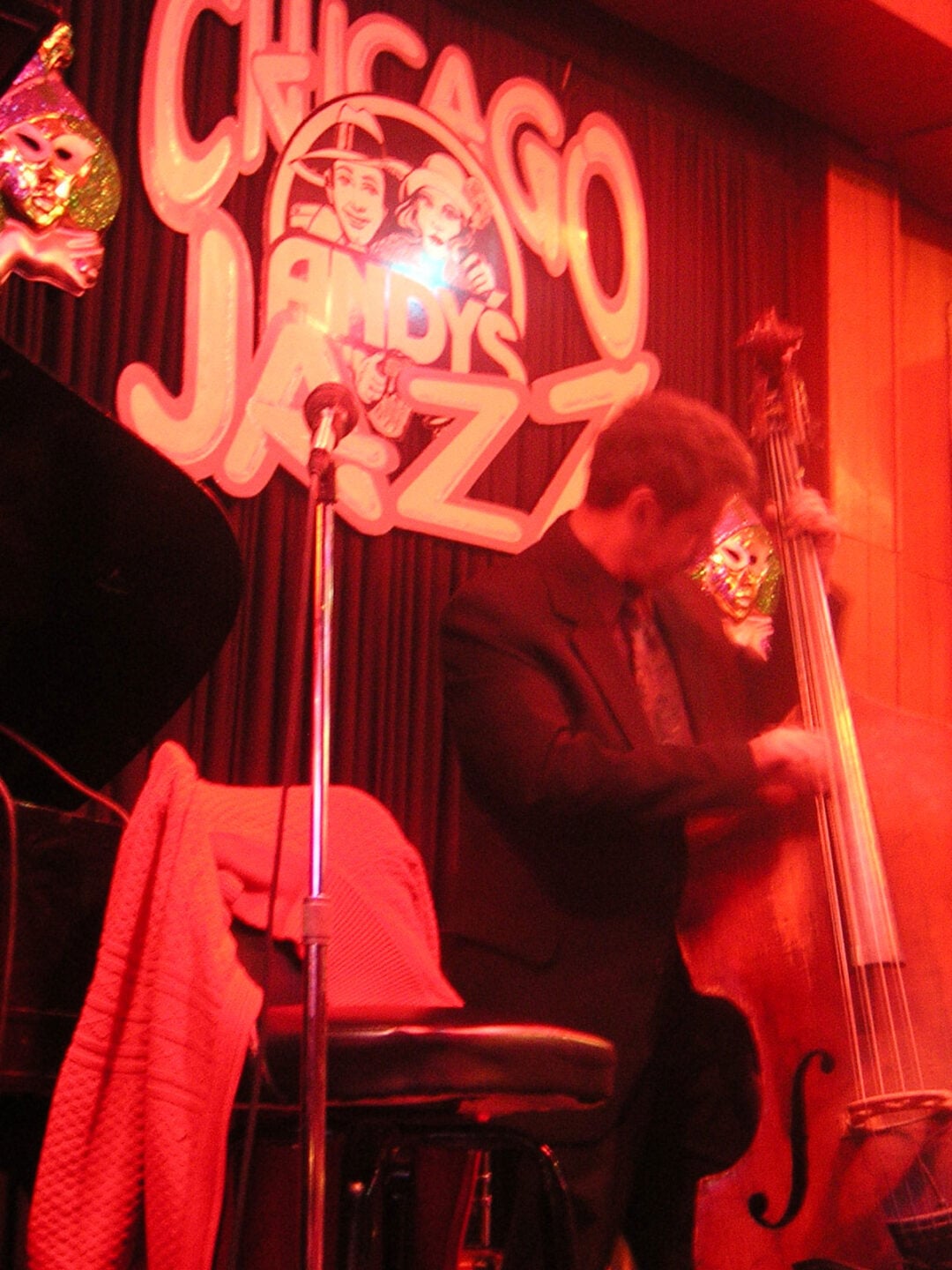 A musician in a suit plays an upright bass onstage at Andy’s Jazz Club in Chicago, with a piano and microphone nearby. Behind him is a neon sign reading “Chicago Andy’s Jazz” and theatrical masks as decoration.