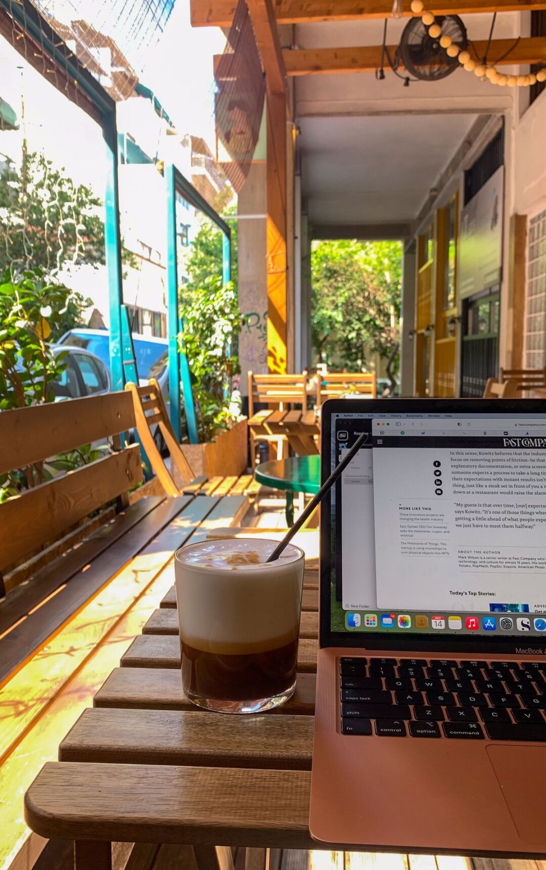 A laptop and a glass of iced coffee with a straw sit on a wooden table at an outdoor café with wooden chairs. Sunlight filters in, plants decorate the space, and a street is visible outside.