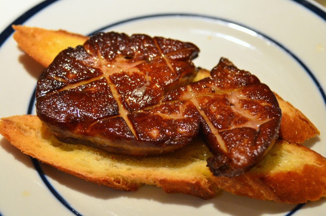 A seared slice of foie gras with a golden-brown crust is placed on top of two toasted baguette slices, served on a white plate with blue trim.