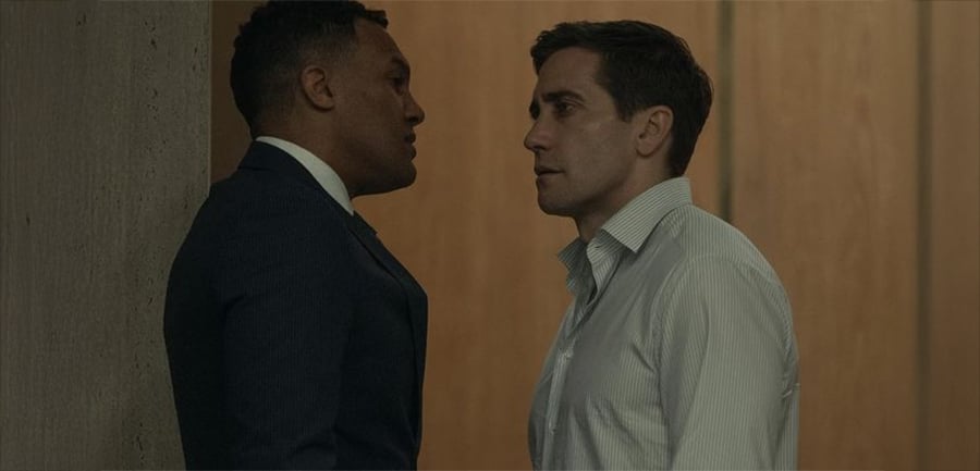 Two men face each other closely in a tense confrontation indoors, one in a dark suit leaning against a wall and the other in a white shirt standing in front of him.