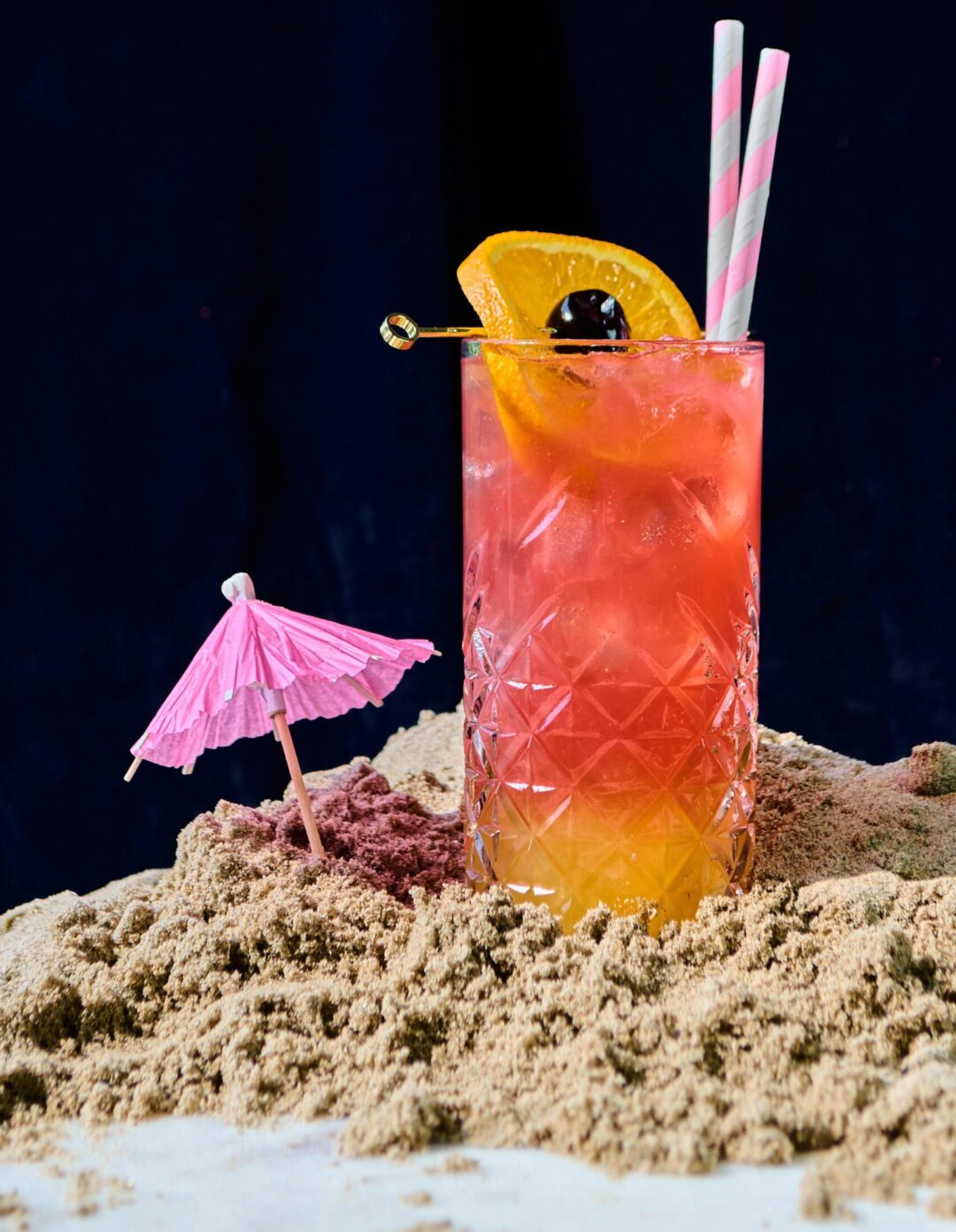 A colorful cocktail garnished with an orange slice and two pink striped straws sits on sand, with a pink paper umbrella nearby, against a dark background.