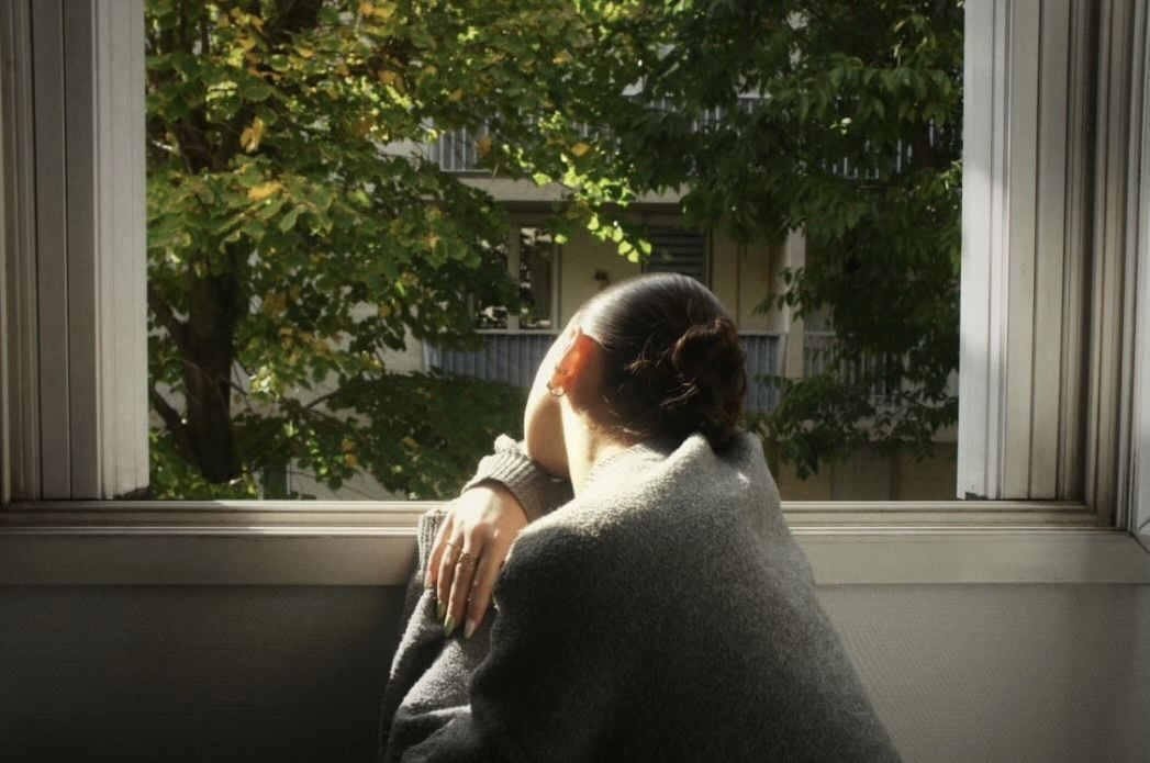 A person wrapped in a gray blanket leans on a windowsill, looking outside at sunlit green trees and an apartment building. Light falls on their face and shoulder, creating a calm and contemplative mood.