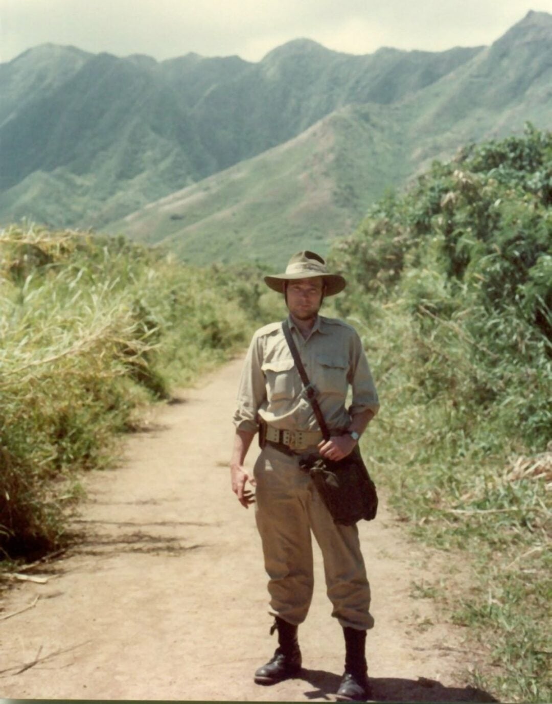 A person in khaki clothing, boots, and a wide-brimmed hat stands on a dirt path surrounded by tall grass, with green mountains in the background under a partly cloudy sky.