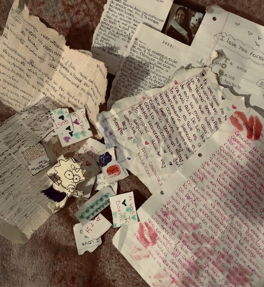A collection of handwritten letters, notes, and drawings scattered on a soft brown surface. Some pages have lipstick kisses, hearts, and doodles. One note features a cartoon drawing, and a small black and white photo is visible.