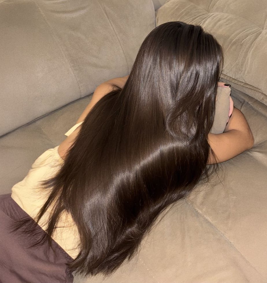 A person with long, straight, shiny dark brown hair lies face down on a beige couch, resting their head on a cushion.