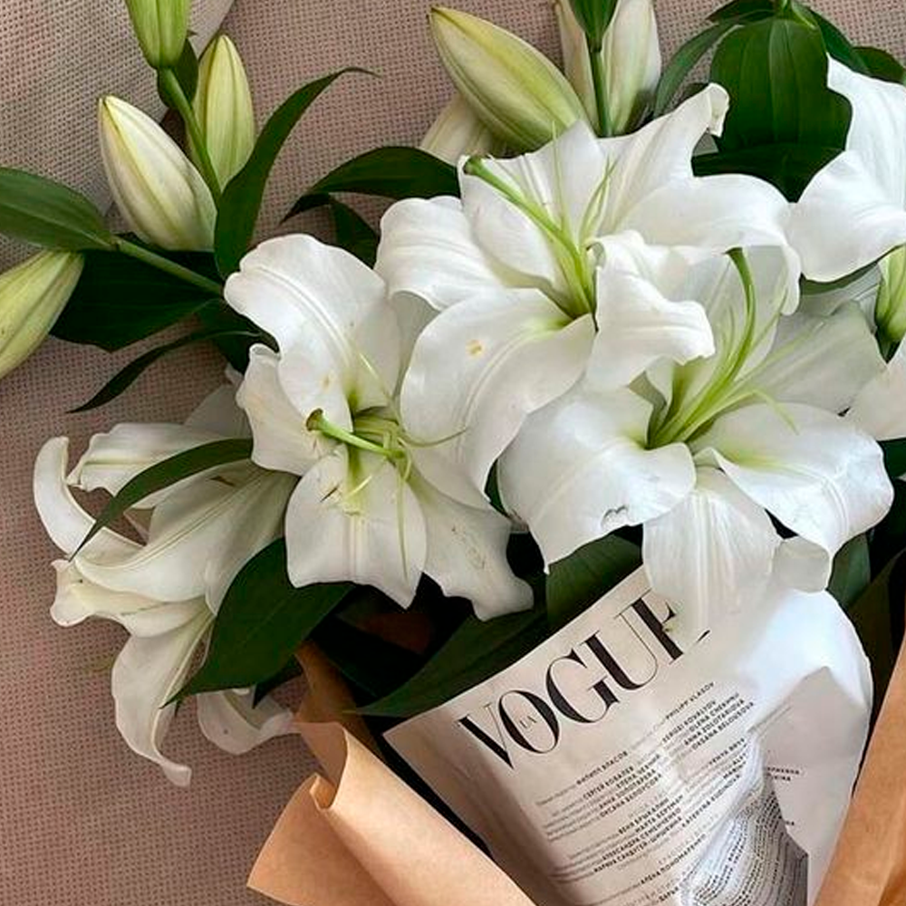 A bouquet of white lilies with green leaves is wrapped in brown paper, with a folded Vogue magazine partially visible among the flowers.
