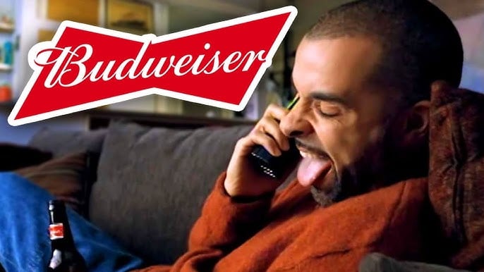 A man sits on a couch holding a Budweiser beer bottle and talking on the phone, sticking out his tongue playfully. The Budweiser logo is prominently displayed in the top left corner.