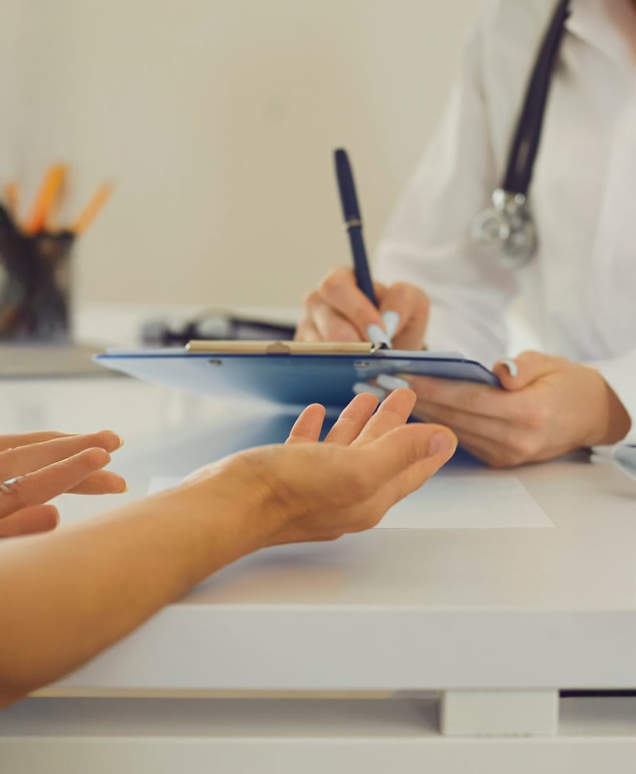 A doctor with a stethoscope writes on a clipboard while sitting at a desk across from a patient, whose hands are extended, during a medical consultation.
