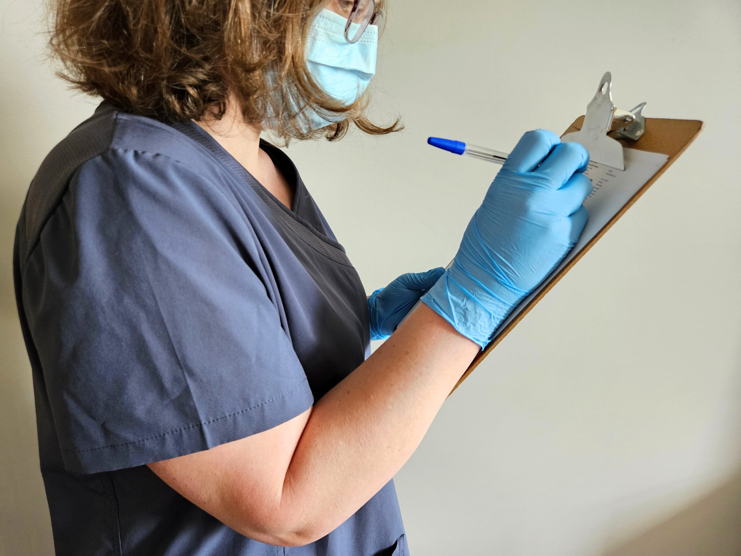 A person wearing blue medical scrubs, gloves, and a face mask writes on a clipboard with a blue pen against a plain background.