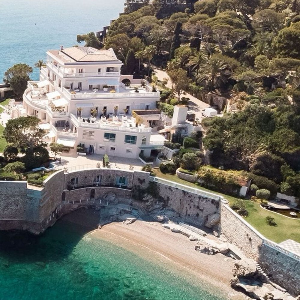 A large, white villa with multiple balconies sits on a seaside cliff, surrounded by lush greenery and overlooking a small, sandy beach with clear turquoise water.