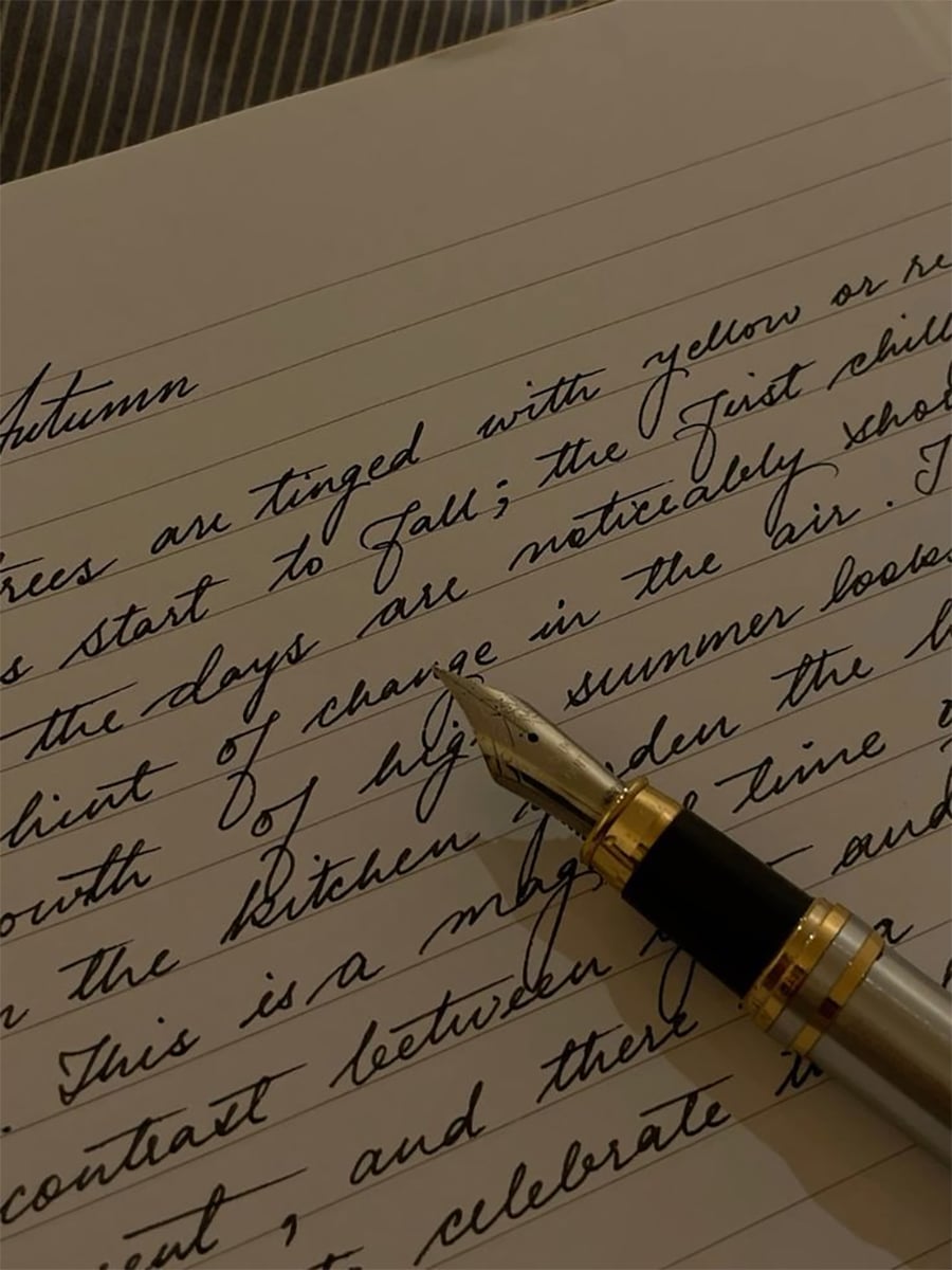 A close-up of a fountain pen resting on an open notebook with neat, cursive handwriting describing the autumn season. The page is lined and the writing is in black ink.