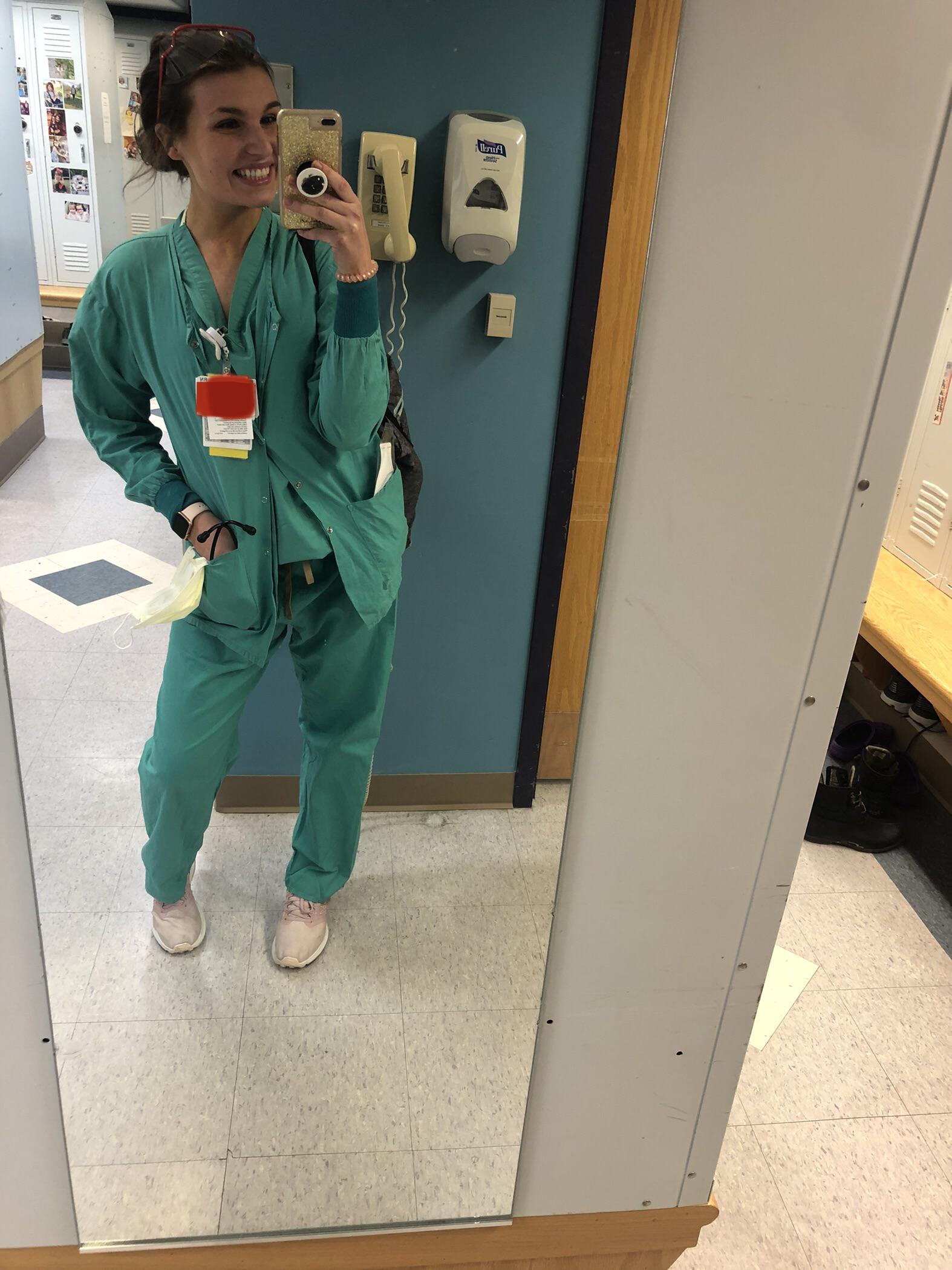 A woman in green medical scrubs and pink shoes smiles and takes a selfie in a mirror inside a hospital locker room. She wears several ID badges and has a pen and stethoscope clipped to her top.