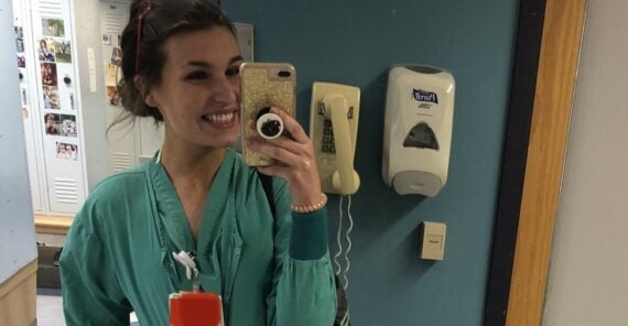 A woman in teal medical scrubs takes a smiling selfie in a mirror, standing in what appears to be a hospital or clinic hallway, with a phone, sanitizer dispenser, and lockers visible in the background.