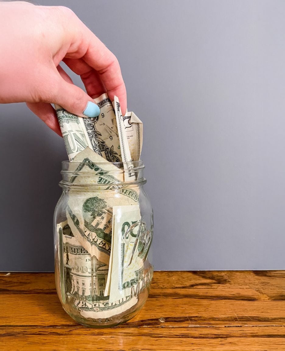 A hand with light blue nail polish places U.S. dollar bills into a glass jar filled with cash, sitting on a wooden surface against a plain gray background.