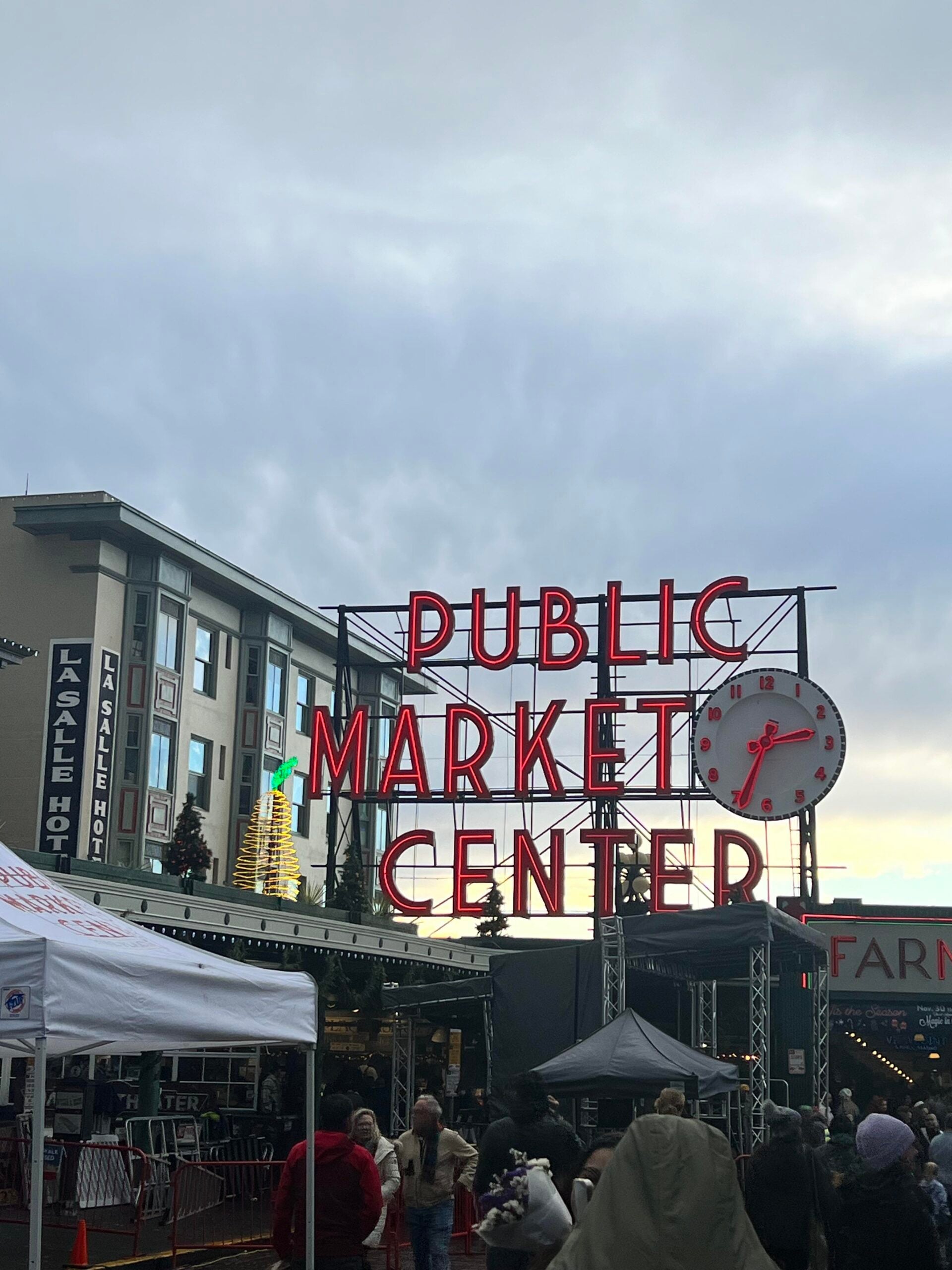 The iconic neon sign and clock of Seattle’s Public Market Center, with buildings in the background, a cloudy sky above, and vendors and crowds gathered beneath tents in the foreground.