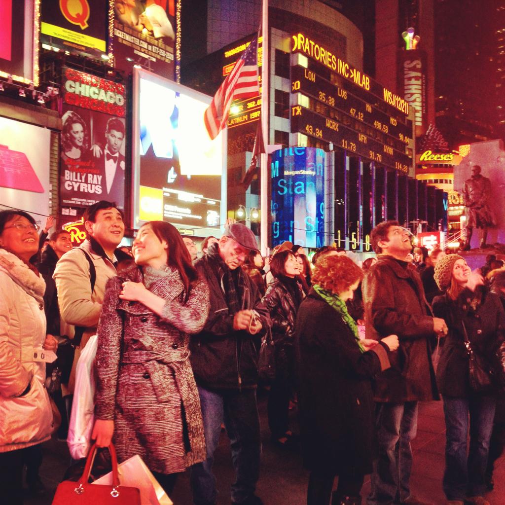 A crowd of people, bundled in winter coats, stands in Times Square at night, surrounded by bright billboards, neon lights, and advertisements. An American flag is visible among the colorful illuminated signs.