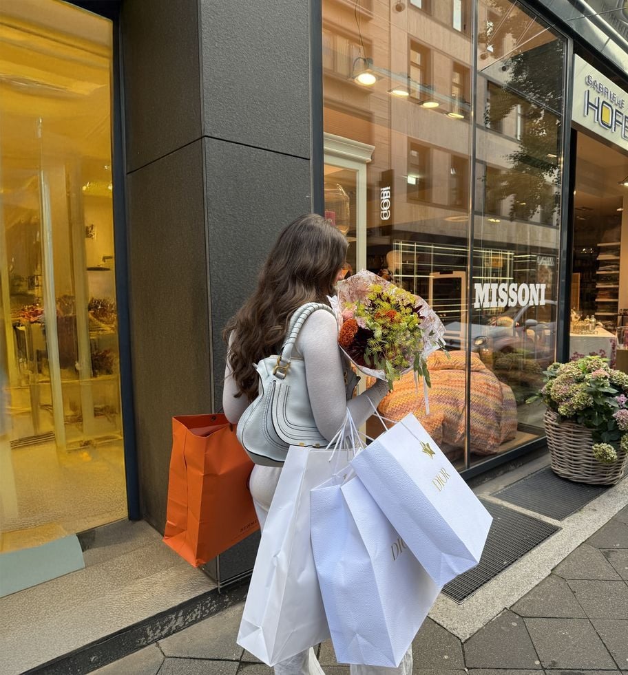 A woman with long brown hair, dressed in white, carries shopping bags and a bouquet of flowers while walking on a city sidewalk past a Missoni store.