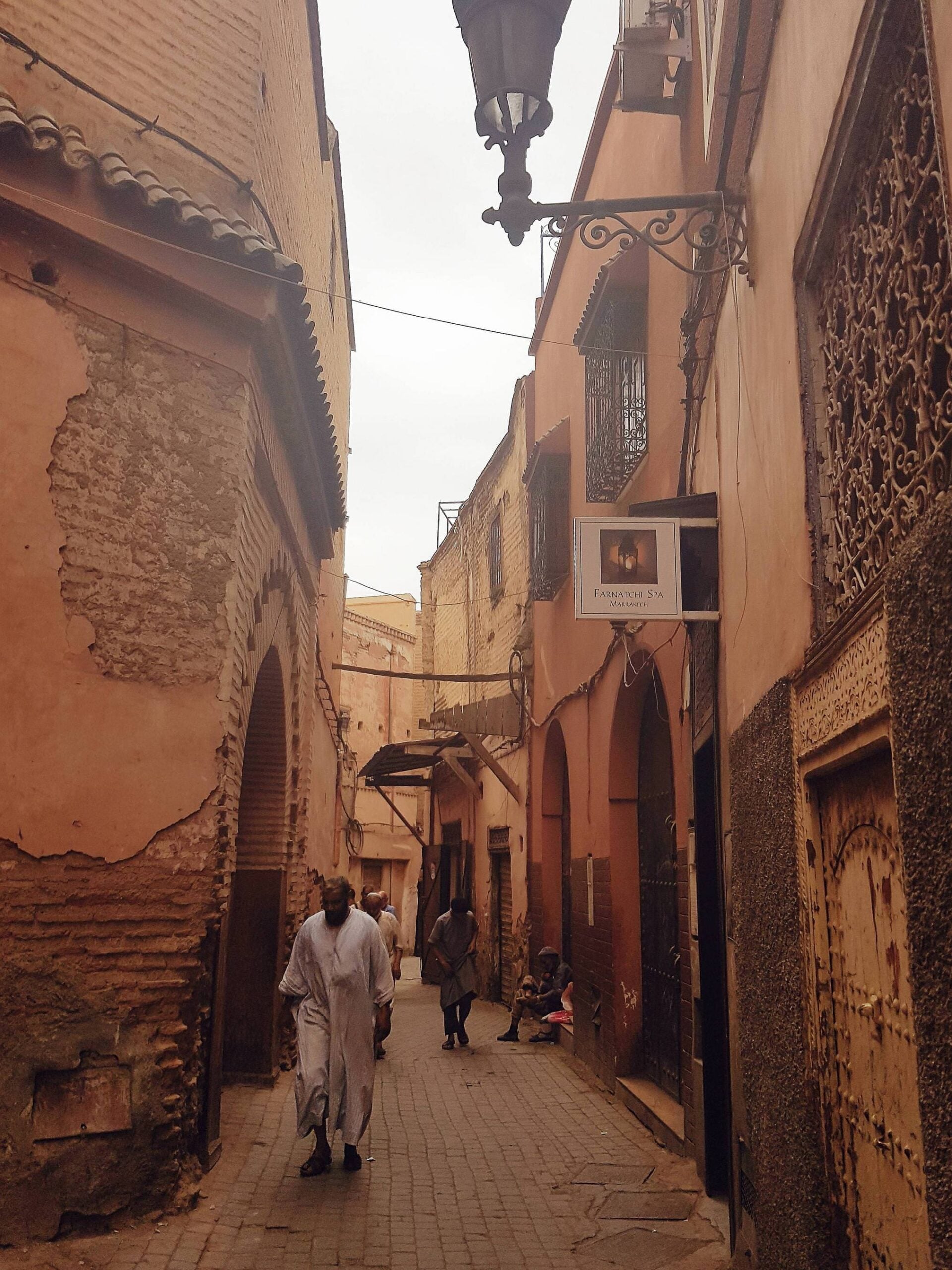 Narrow, winding street in an old city with pink and brown buildings, decorative metalwork, a lantern, and people walking and sitting along the cobblestone path under a cloudy sky.