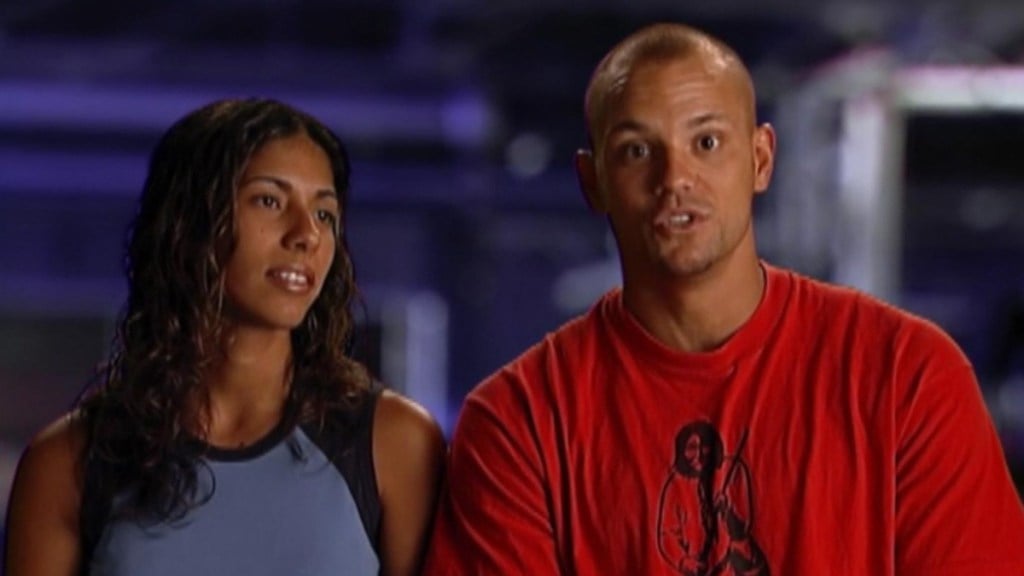 A woman with long curly hair and a sleeveless blue top sits next to a man with short hair wearing a bright red t-shirt, both looking slightly off camera against a blurred dark background.