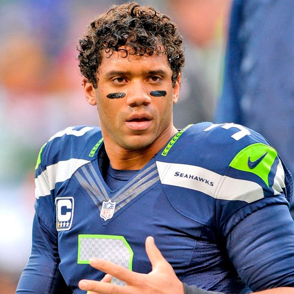 A football player in a Seattle Seahawks uniform with black eye paint stands on the field, looking focused. The uniform displays the Seahawks logo and captain's patch.