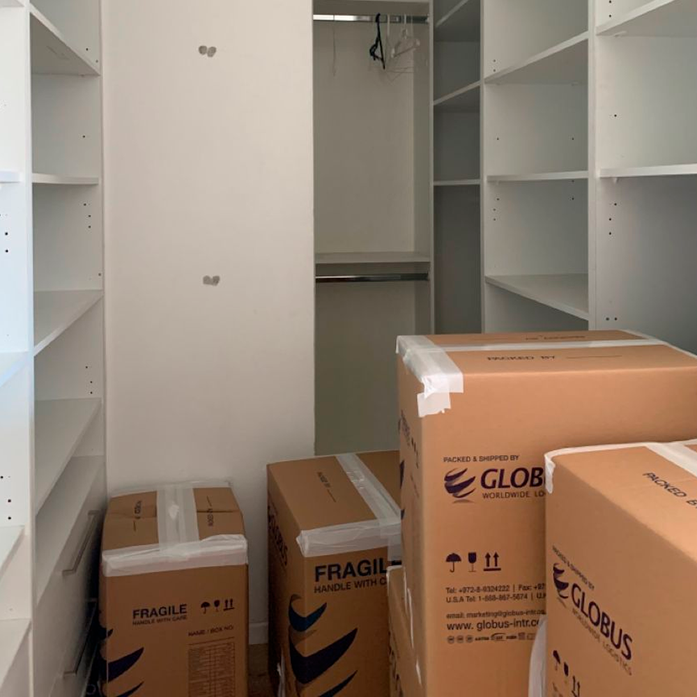 A walk-in closet with empty white shelves and several large, sealed moving boxes labeled "Globus" on the floor, suggesting someone is packing or moving in.