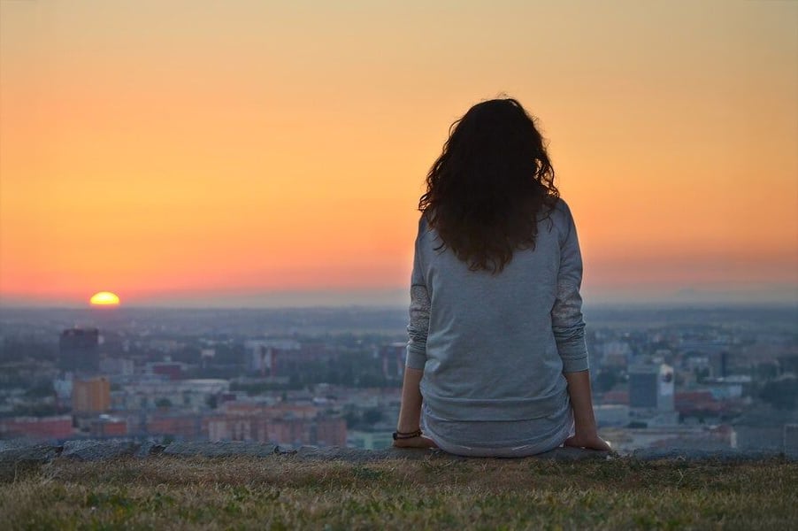A person with long, curly hair sits on grass facing a cityscape, watching the sun set over the horizon with an orange and yellow sky.